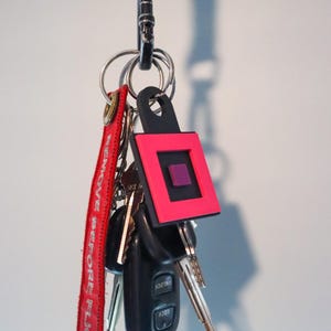 K&ouml;nnte beinhalten: Ein Schl&uuml;sselbund, der an einem schwarzen Metallkarabiner befestigt ist. Die Schl&uuml;ssel umfassen einen Autoschl&uuml;sselanh&auml;nger und mehrere Metallschl&uuml;ssel. Ein rotes Etikett mit dem Text "REMOVE BEFORE FLIGHT" ist angebracht. Ein schwarzer und pinkfarbener Schl&uuml;sselanh&auml;nger ist ebenfalls vorhanden.