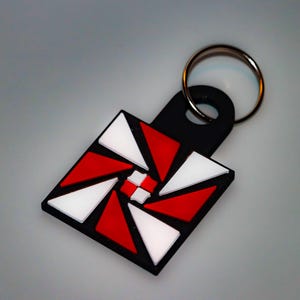 May include: Black keychain with a square pendant featuring a red, white, and black geometric design. The design is a series of triangles arranged in a spiral pattern. The keychain has a silver metal ring.