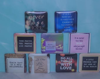 Könnte beinhalten: Eine Sammlung von neun quadratischen Magneten mit verschiedenen motivierenden Zitaten und Sprüchen wie never lose hope, think positive, do all things with love und each day is a new beginning, angeordnet in einer Pyramidenform auf blauem Grund.
