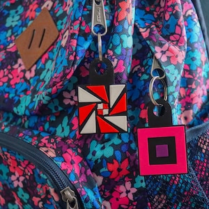 K&ouml;nnte beinhalten: Zwei geometrische Schl&uuml;sselanh&auml;nger h&auml;ngen an einem Rei&szlig;verschluss eines Rucksacks mit Blumenmuster. Einer hat ein rotes, wei&szlig;es und schwarzes Design, der andere ist ein rosa Quadrat mit einem schwarzen und einem violetten Quadrat.