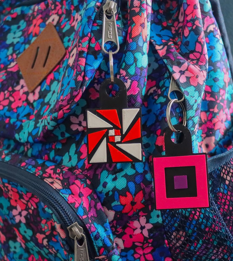 K&ouml;nnte beinhalten: Zwei geometrische Schl&uuml;sselanh&auml;nger h&auml;ngen an einem Rei&szlig;verschluss eines Rucksacks mit Blumenmuster. Einer hat ein rotes, wei&szlig;es und schwarzes Design, der andere ist ein rosa Quadrat mit einem schwarzen und einem violetten Quadrat.