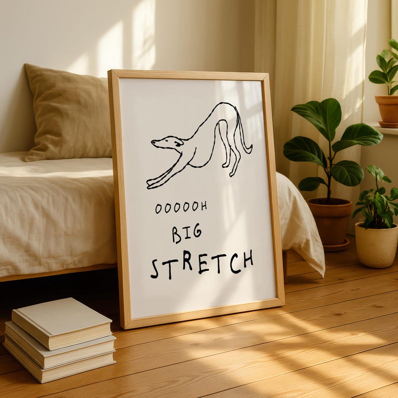 A framed poster with a cartoon dog and the text "OODDOH BIG STRRETCH" is displayed on a wooden floor in a room with a bed, potted plants, and books.