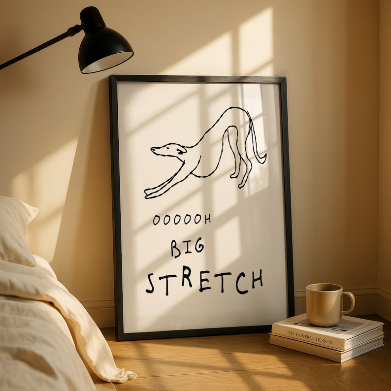 a framed poster with a simple black and white line drawing of a dog, accompanied by the text "OOOOH BIG STRETCH" in bold letters. The poster is displayed on a wall next to a bed, with a lamp and a stack of books visible in the background.