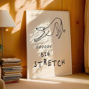A framed poster with a simple black and white drawing of a dog and the text "OOOH BIG STRETCH" on it, placed on a wooden floor next to a stack of books and a lamp.