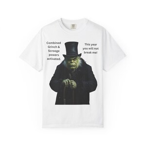 May include: White t-shirt featuring a combined Grinch and Scrooge graphic. The image shows a green-skinned character in a black coat and top hat, holding a cane. Text on the shirt reads "Combined Grinch & Scrooge powers activated. This year you will not break me!"