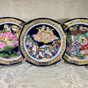 May include: Three decorative plates with intricate designs. Each plate features a dark blue background with a gold filigree border. The center of each plate showcases a colorful, detailed illustration of a religious scene. The plates are displayed on clear stands.