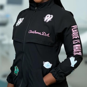 May include: Black zip-up jacket with dental-themed patches and text. The jacket features a smiling tooth graphic, a mask, and the words "Plaque is whack" on the sleeve. The name "Chrishona, D.A." is embroidered in pink.