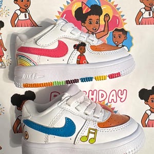 Op de afbeelding: Witte peutersneakers met kleurrijke accenten en aangepaste illustraties. Eén schoen heeft een roze Nike swoosh en een regenboogontwerp, de andere een blauwe swoosh en muzieknoten. Het woord "BIRTHDAY" is zichtbaar.