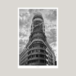 May include: A black and white architectural photograph of the Edificio Carrión in Madrid, featuring the prominent Schweppes sign on its curved facade. The building displays multiple levels of windows under a cloudy sky, captured from a low angle.