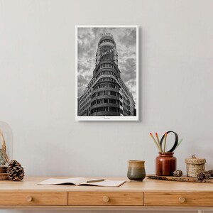 May include: A black and white framed wall art print featuring the iconic Edificio Carrión in Madrid, showcasing its curved facade, multiple rows of windows, and the prominent illuminated Schweppes and Capitol signs against a cloudy sky.