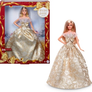 May include: A blonde Barbie doll wearing a gold and white patterned ball gown with a sheer neckline and bow. The doll has gold earrings and is in a red and gold box labeled "Holiday Barbie 2025".