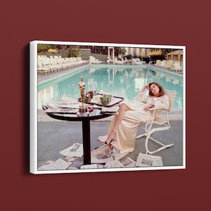 May include: A framed photograph depicts a woman relaxing by a pool. She is seated in a white chair, wearing a long, light-colored dress and high heels. A small table holds an Oscar statuette, a tray with a cup, and newspapers. The pool is in the background.