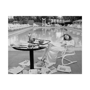May include: Black and white photograph of a person relaxing by a pool. The person is seated in a white chair, wearing a long satin dress and heels. A table with an Oscar statue and a tray of items sits nearby, surrounded by newspapers.