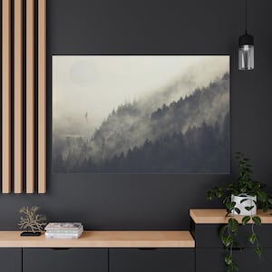 May include: A canvas print depicting a misty mountain scene with a large, pale sun. The artwork features a dense forest on a hillside, partially obscured by fog. A small building and a bird are visible.