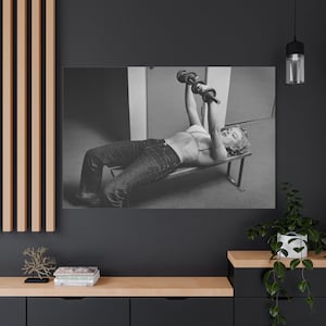 May include: Black and white print of a woman lifting weights while lying on a bench. She is wearing jeans and a bra, holding two dumbbells above her chest. The image is a vintage style photograph.