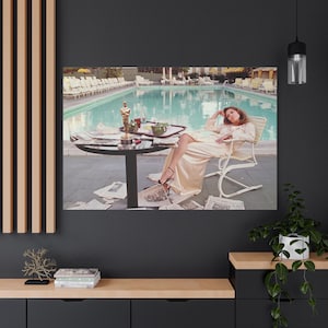 May include: A canvas print depicts a woman relaxing by a pool. She is wearing a cream-colored gown and sitting in a white chair. An Oscar statue sits on a table with newspapers and a tray of items. The pool is in the background.