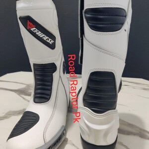 Puede incluir: Par de botas de moto blancas y negras con el logo Dainese. Las botas tienen detalles negros y una placa plateada en la puntera. El texto "Road Raptor Pk" es visible en el lateral. Botas de motorista.