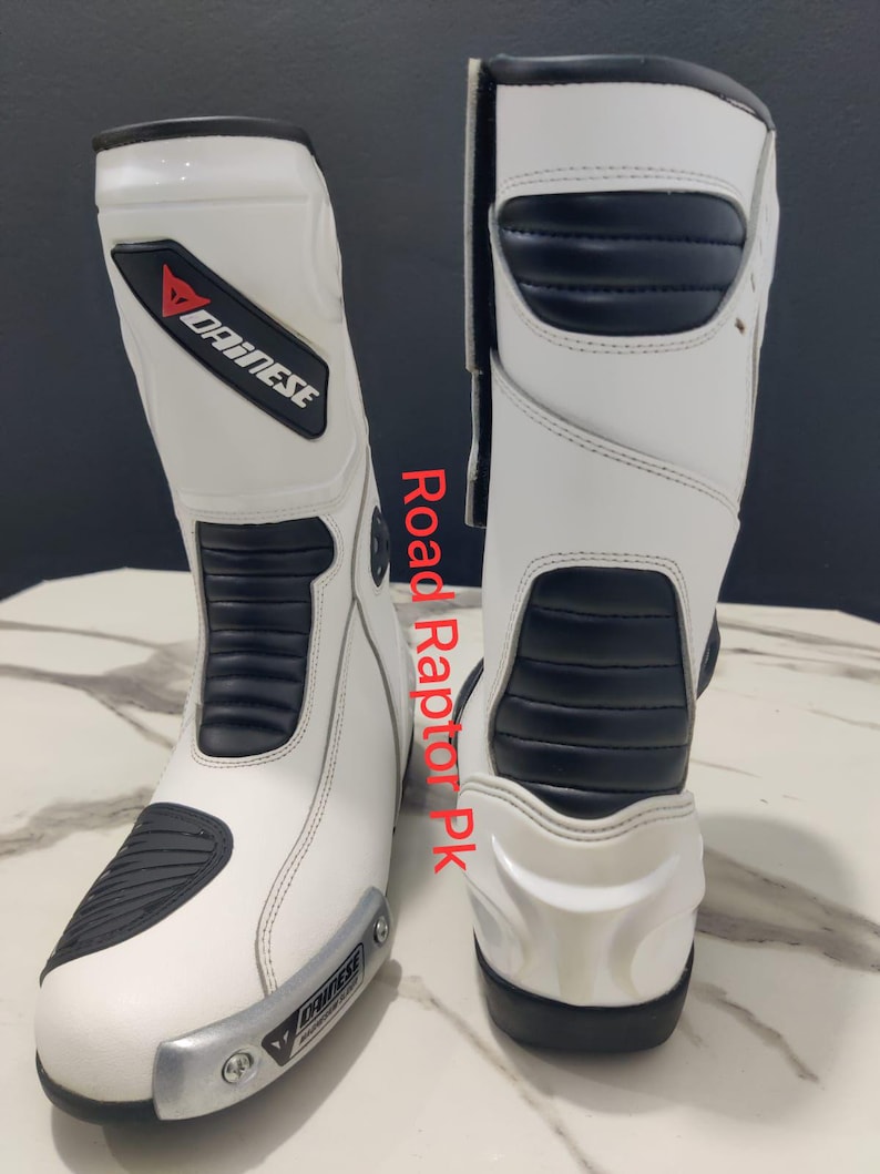 Puede incluir: Par de botas de moto blancas y negras con el logo Dainese. Las botas tienen detalles negros y una placa plateada en la puntera. El texto "Road Raptor Pk" es visible en el lateral. Botas de motorista.