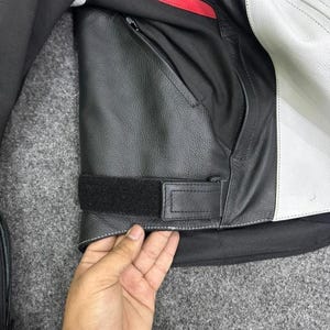 Puede incluir: Primer plano de una chaqueta de moto de cuero negro y blanco. La chaqueta presenta un exterior de cuero negro con un bolsillo con cremallera, una correa de velcro y costuras blancas. Una franja roja acent&uacute;a la zona de los hombros.