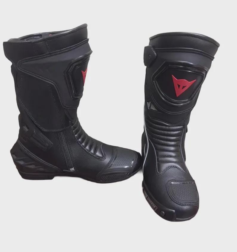 May include: Pair of black motorcycle boots with red accents. The boots have a tall design, a zipper closure, and a reinforced toe. The boots feature a red logo on the side and are designed for protection and performance.