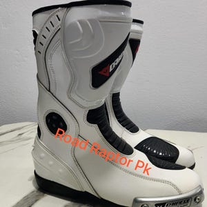 Puede incluir: Par de botas de moto blancas y negras con la marca "DAINESE" visible. Las botas tienen un dise&ntilde;o elegante con elementos de protecci&oacute;n, incluido un deslizador de magnesio. El texto "Road Raptor Pk" tambi&eacute;n es visible.