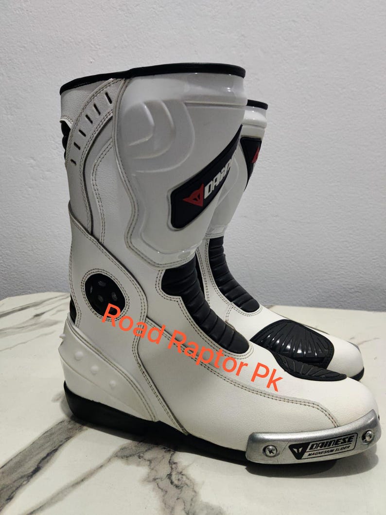 Puede incluir: Par de botas de moto blancas y negras con la marca "DAINESE" visible. Las botas tienen un dise&ntilde;o elegante con elementos de protecci&oacute;n, incluido un deslizador de magnesio. El texto "Road Raptor Pk" tambi&eacute;n es visible.