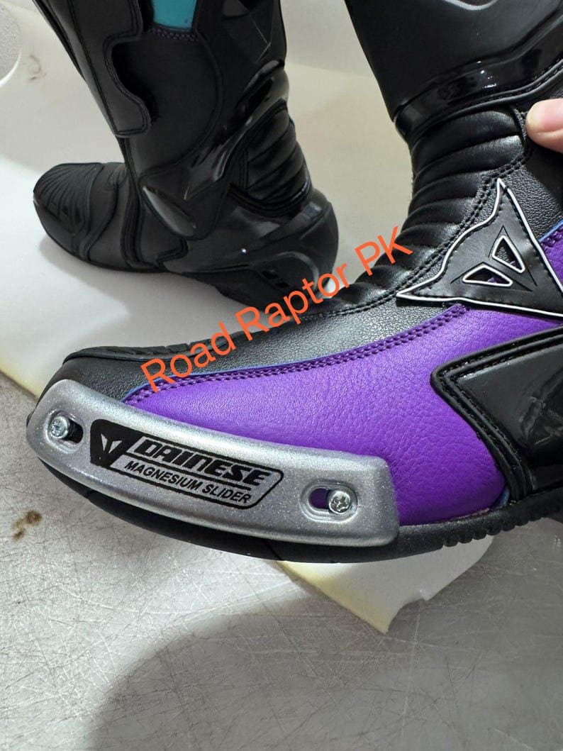 Może przedstawiać: Czarne i fioletowe buty motocyklowe ze srebrnym ochraniaczem palc&oacute;w. Buty mają markę "DAINESE". Zaprojektowane do jazdy na motocyklu, zapewniają ochronę i styl.