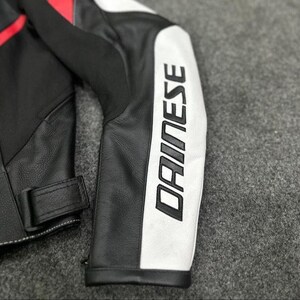 Puede incluir: Primer plano de una manga de chaqueta de moto de cuero negro y blanco. La manga presenta el nombre de la marca "DAINESE" en letras blancas grandes y en negrita. Se ven detalles en rojo.
