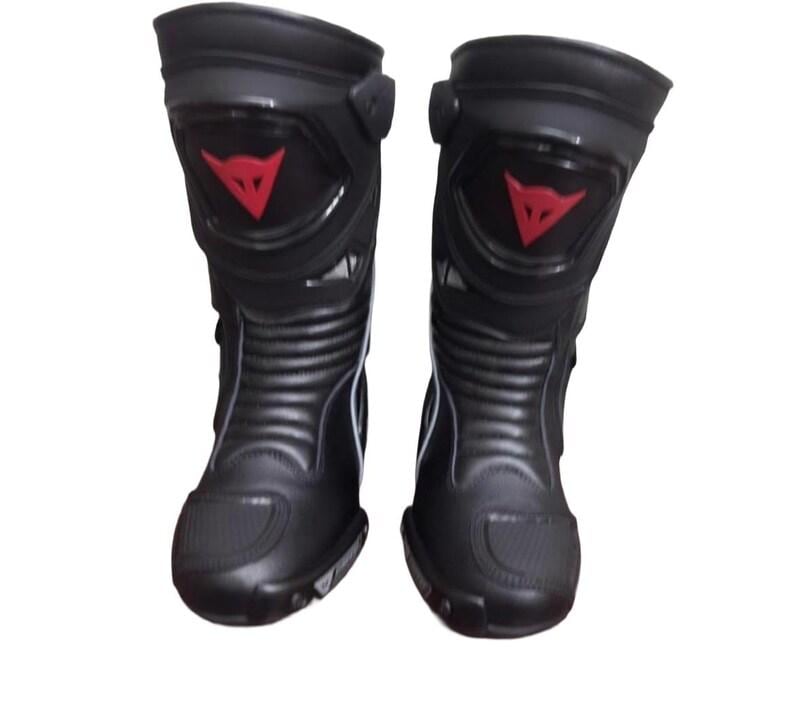 May include: Pair of black motorcycle boots with a red logo on the shin. The boots have a sleek design with reinforced areas and a ridged lower section. The boots are designed for protection and performance.