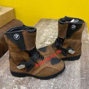 Puede incluir: Un par de botas de moto de cuero marrón con detalles en negro, una correa con hebilla ajustable negra en el empeine y un emblema circular de BMW en el cuello superior. Las botas tienen una suela negra y paneles protectores negros.