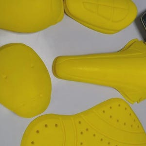 Puede incluir: Equipo de protecci&oacute;n para motocicletas de color amarillo brillante, que incluye protectores de hombros y espalda. Las piezas tienen varias formas y tama&ntilde;os, algunas con orificios de ventilaci&oacute;n. La imagen tambi&eacute;n muestra una revista con las palabras "SMOOTH WAYS".