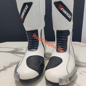 Puede incluir: Par de botas de moto blancas con detalles negros y el logotipo "Dainese". Las botas presentan un dise&ntilde;o elegante con elementos de protecci&oacute;n, incluyendo refuerzo en la puntera y el tobillo. El texto "Road Raptor Pk" es visible.