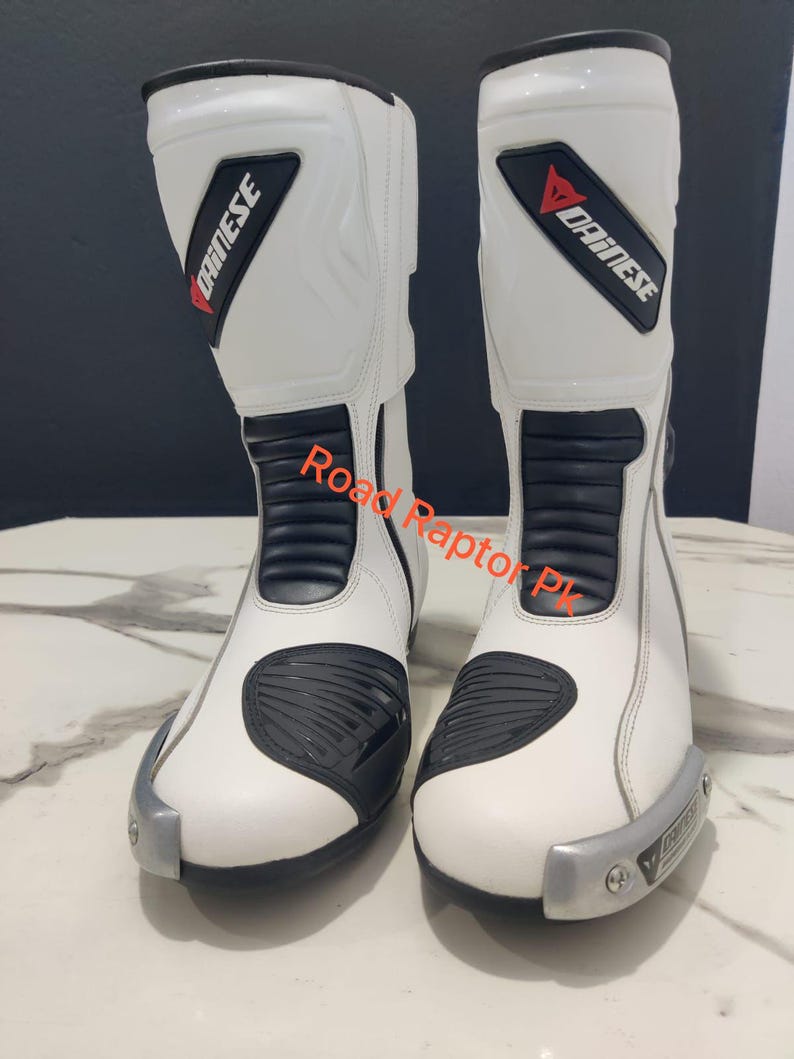 Puede incluir: Par de botas de moto blancas con detalles negros y el logotipo "Dainese". Las botas presentan un dise&ntilde;o elegante con elementos de protecci&oacute;n, incluyendo refuerzo en la puntera y el tobillo. El texto "Road Raptor Pk" es visible.