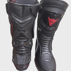 May include: Pair of black motorcycle boots with protective features. The boots have a high-top design, with reinforced panels and a red logo on the side. The boots are made of a durable material and are designed for safety and performance.