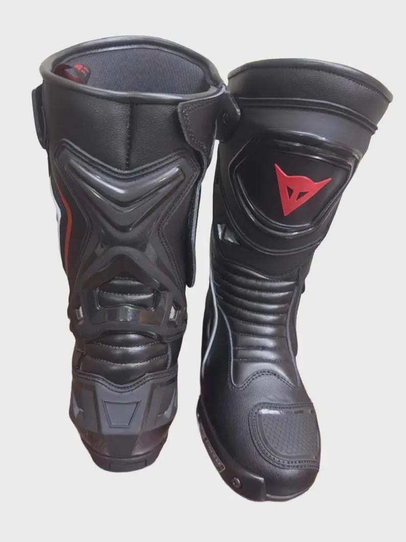May include: Pair of black motorcycle boots with protective features. The boots have a high-top design, with reinforced panels and a red logo on the side. The boots are made of a durable material and are designed for safety and performance.