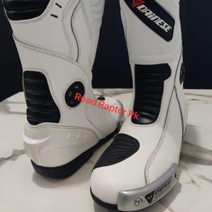 Puede incluir: Par de botas de moto blancas con detalles negros y el logotipo "DAINESE". Las botas tienen un dise&ntilde;o elegante con &aacute;reas reforzadas para protecci&oacute;n. Las botas son de cuero y tienen una placa met&aacute;lica plateada en la parte delantera.