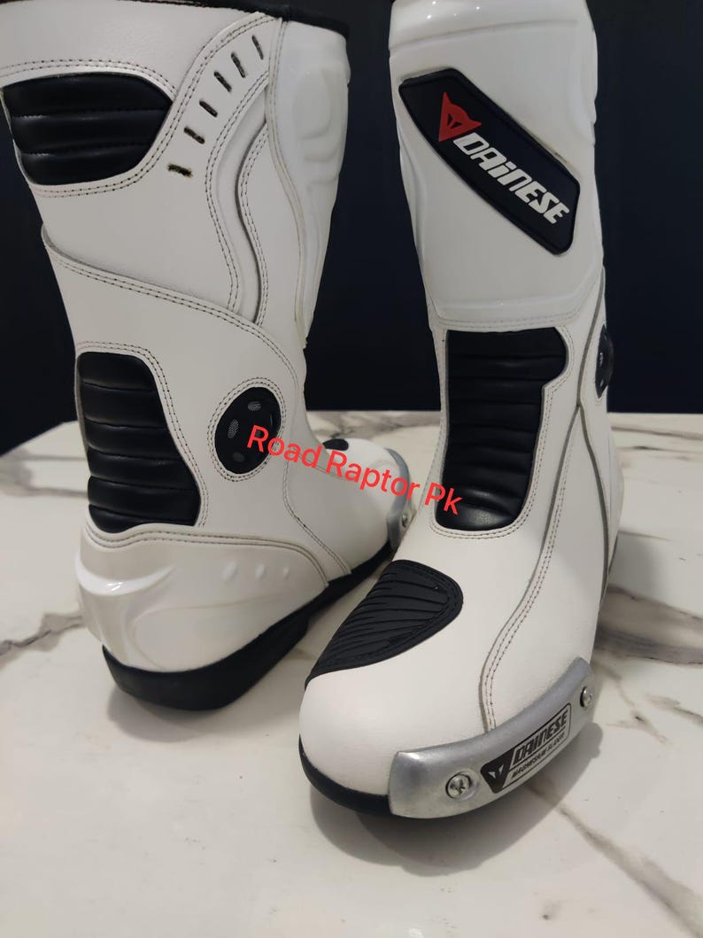 Puede incluir: Par de botas de moto blancas con detalles negros y el logotipo "DAINESE". Las botas tienen un dise&ntilde;o elegante con &aacute;reas reforzadas para protecci&oacute;n. Las botas son de cuero y tienen una placa met&aacute;lica plateada en la parte delantera.