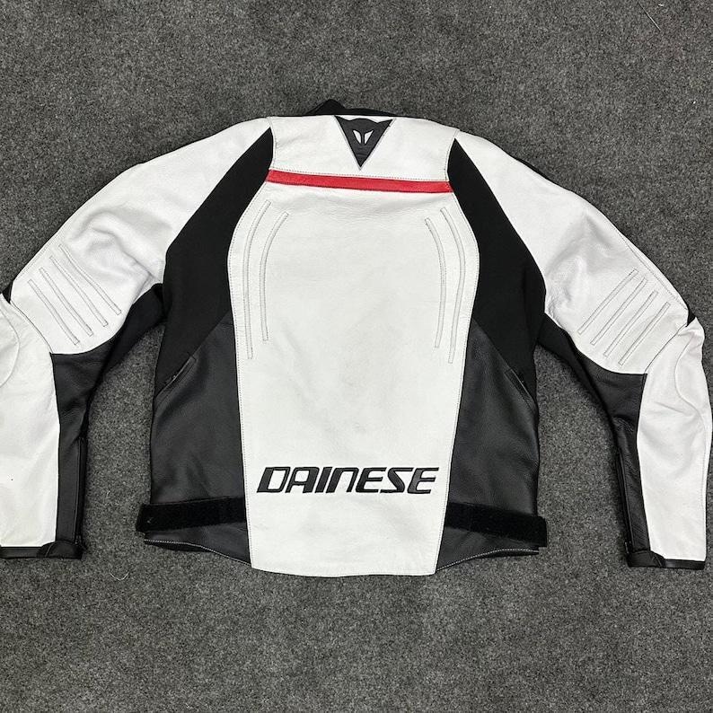 Puede incluir: Chaqueta de moto blanca y negra con detalles en rojo. La chaqueta presenta el logotipo "DAINESE" en letras blancas. Paneles negros en los laterales y mangas, con paneles blancos en la parte delantera y trasera. Chaqueta de motorista.