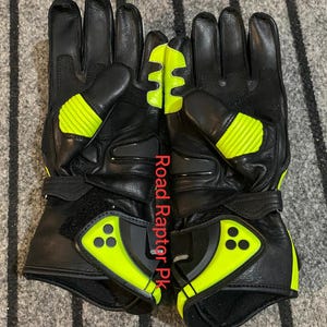 May include: Pair of black motorcycle gloves with neon yellow accents. The gloves feature reinforced knuckles and a velcro wrist strap. The text "Road Raptor Pk" is printed vertically on the gloves.