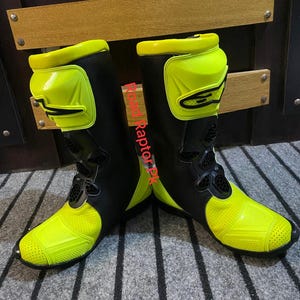 May include: Pair of black and neon yellow motorcycle boots. The boots have a high-top design with protective panels on the shin and ankle. The toe and heel areas are reinforced. The boots are designed for safety and performance.