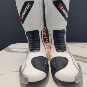 Puede incluir: Par de botas de moto blancas con detalles negros y el logotipo "DAINESE" en rojo y negro. Las botas tienen elementos de protecci&oacute;n y una puntera plateada. El texto "Road Raptor PK" est&aacute; impreso en el lateral.
