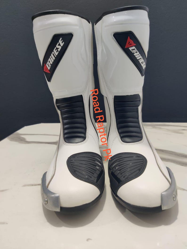 Puede incluir: Par de botas de moto blancas con detalles negros y el logotipo "DAINESE" en rojo y negro. Las botas tienen elementos de protecci&oacute;n y una puntera plateada. El texto "Road Raptor PK" est&aacute; impreso en el lateral.
