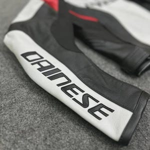 Puede incluir: Primer plano de una manga de chaqueta de moto de cuero blanco y negro. La palabra "DAINESSE" est&aacute; impresa en letras negras grandes sobre una franja blanca. La chaqueta parece estar hecha de cuero de alta calidad.