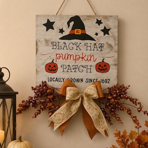 Può includere: Un cartello quadrato in legno con finitura bianca anticata, raffigurante un cappello da strega nero, due zucche arancioni e stelle nere. Il testo recita: BLACK HAT PUMPKIN PATCH LOCALLY GROWN SINCE 1692. È decorato con un grande fiocco beige e dorato.