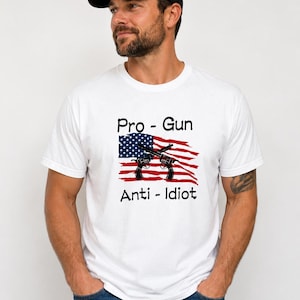 Peut inclure: Un t-shirt blanc à manches courtes avec un graphique du drapeau américain et deux revolvers noirs croisés superposés. Le texte Pro - Gun est imprimé au-dessus du drapeau et le texte Anti - Idiot est imprimé en dessous en noir.