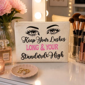 Könnte beinhalten: Eine rechteckige Reißverschlusstasche aus cremefarbenem Canvas mit einer schwarzen Grafik von zwei Augen mit langen Wimpern und dem schwarzen und pinken Text: Keep Your Lashes LONG & YOUR Standards High. Die Tasche hat einen goldfarbenen Metallreißverschluss.