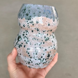 May include: A handmade ceramic vase with a unique speckled design. The vase features a light blue top, a speckled white and tan middle section, and a rounded shape. Dark green speckles are scattered across the surface.