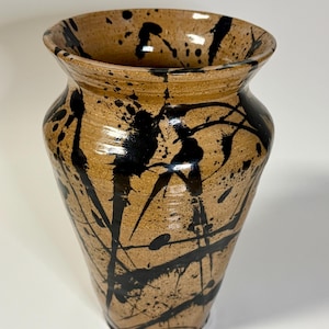 May include: A ceramic vase with a tan, speckled base featuring a bold black paint splatter design. The vessel has a wide, flared rim that tapers down to a narrower base, showcasing a glossy finish over the textured, earthy clay surface.