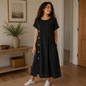 May include: A black midi-length dress with short sleeves, a round neckline, and a vertical row of light-colored buttons running down the front right side. The dress features a relaxed fit with a subtle gathered detail at the waist and white sneakers.