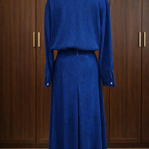 May include: A long, royal blue dress displayed on a mannequin. The dress features a collared, button-down top with long sleeves and a matching floor-length skirt. The fabric has a textured appearance. The dress is displayed against a backdrop of wooden cabinets.